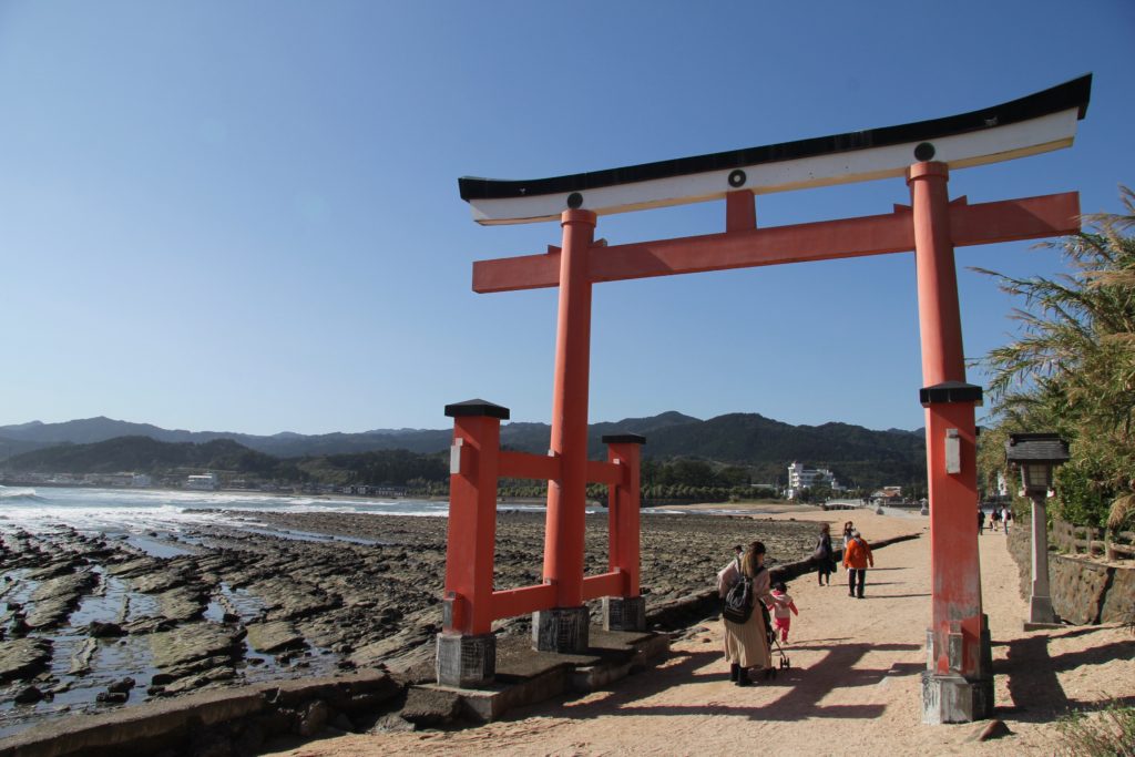 torii gate on a small island in japan, with a few people strolling through, the shore in background